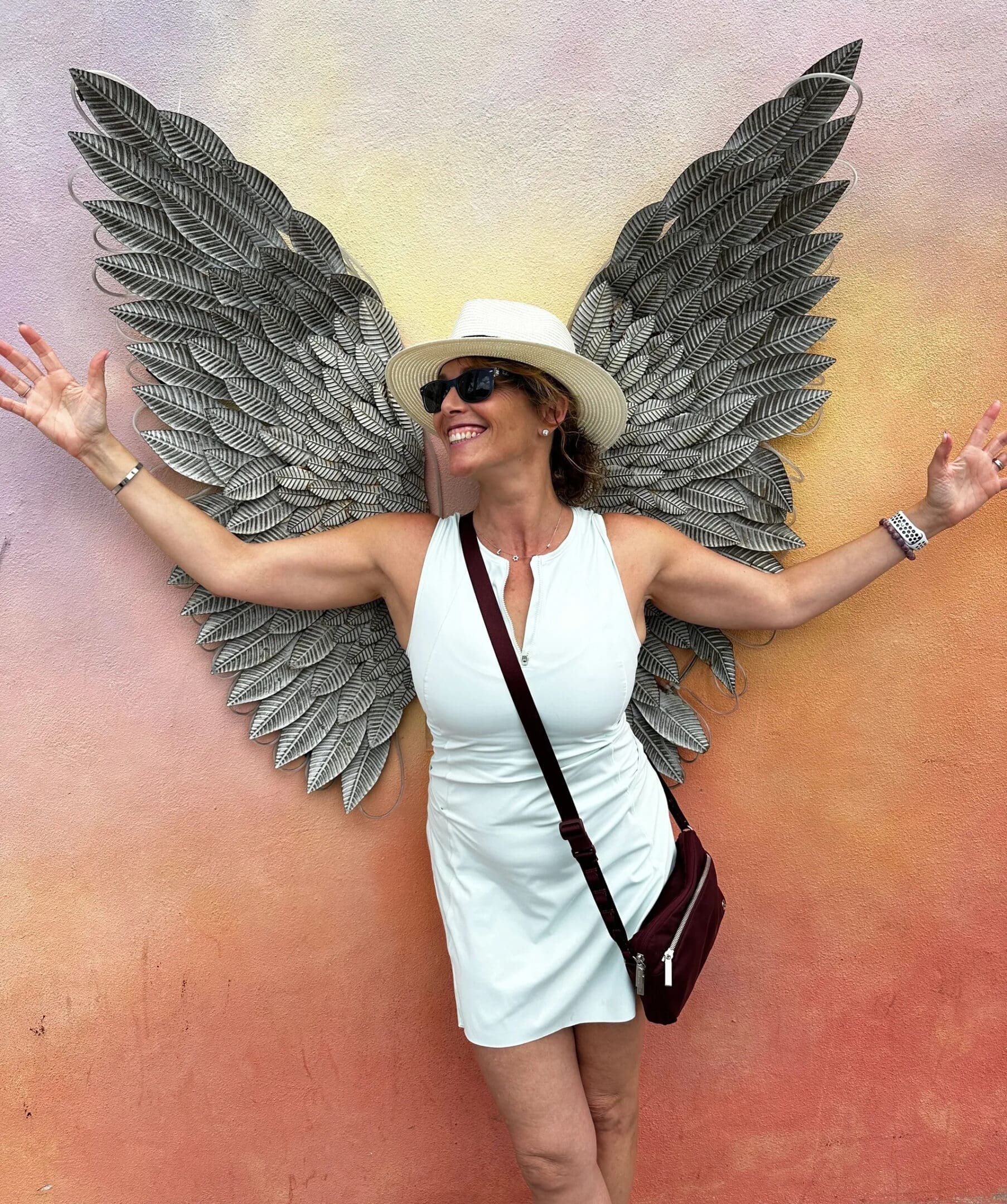 Woman posing with angel wings mural, wearing a white dress and hat.