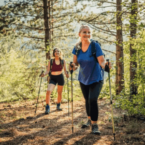Two women hiking with trekking poles on a sunny forest trail.