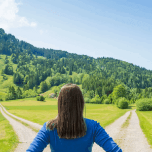 A woman with a blue jacket stands at a fork in a rural path surrounded by green hills.