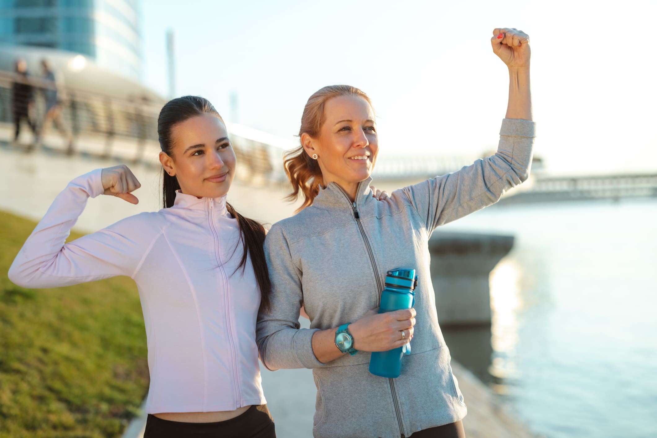 Two female friends celebrate fitness outdoors, flexing their arms and smiling after a morning workout by the waterfront. One holds a reusable water bottle while both wear athletic jackets and leggings.