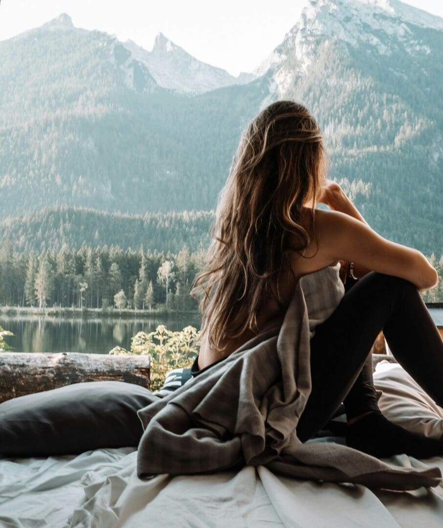Woman sitting up in bed at dawn looking out at mountains and a still lake