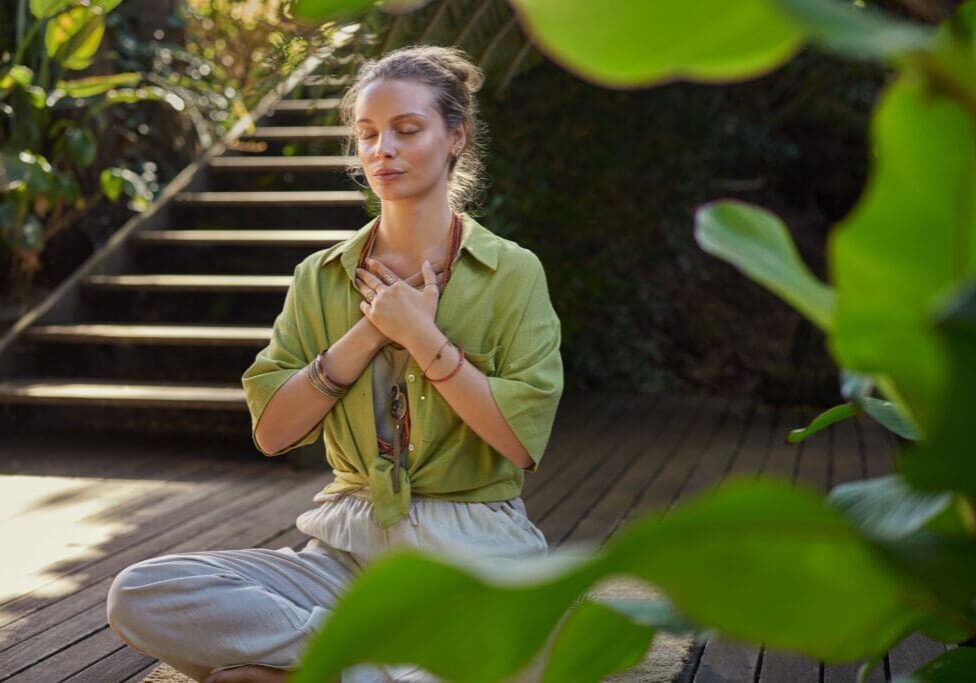 Peaceful woman sits cross-legged in a meditative state, hands over her heart, expressing gratitude and mindfulness in a lush. Girl connects with her emotions during a reflective moment, embracing inner peace. Calm woman practices a heart-centered meditation, cultivating self-love and healing energy, surrounded by greenery.