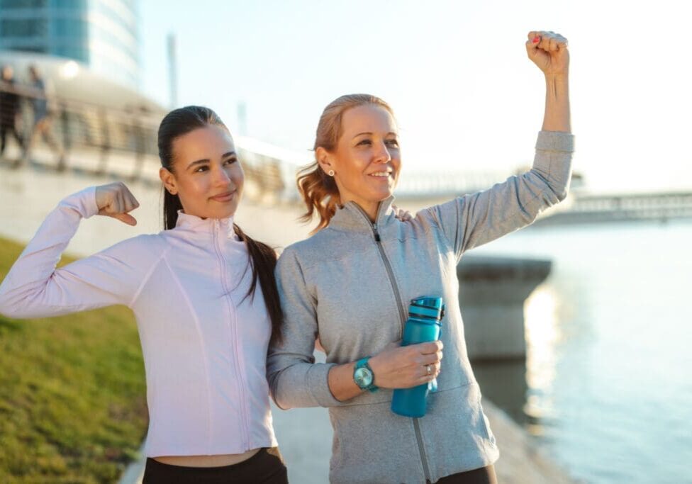 Two female friends celebrate fitness outdoors, flexing their arms and smiling after a morning workout by the waterfront. One holds a reusable water bottle while both wear athletic jackets and leggings.